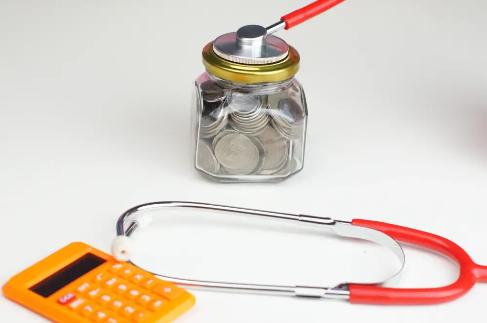 Cofre de vidro cheio de moedas com estetoscópio apoiado e uma calculadora ao lado, simbolizando a relação entre custos em saúde, sinistralidade e bem-estar corporativo. Sinistralidade de plano de saúde