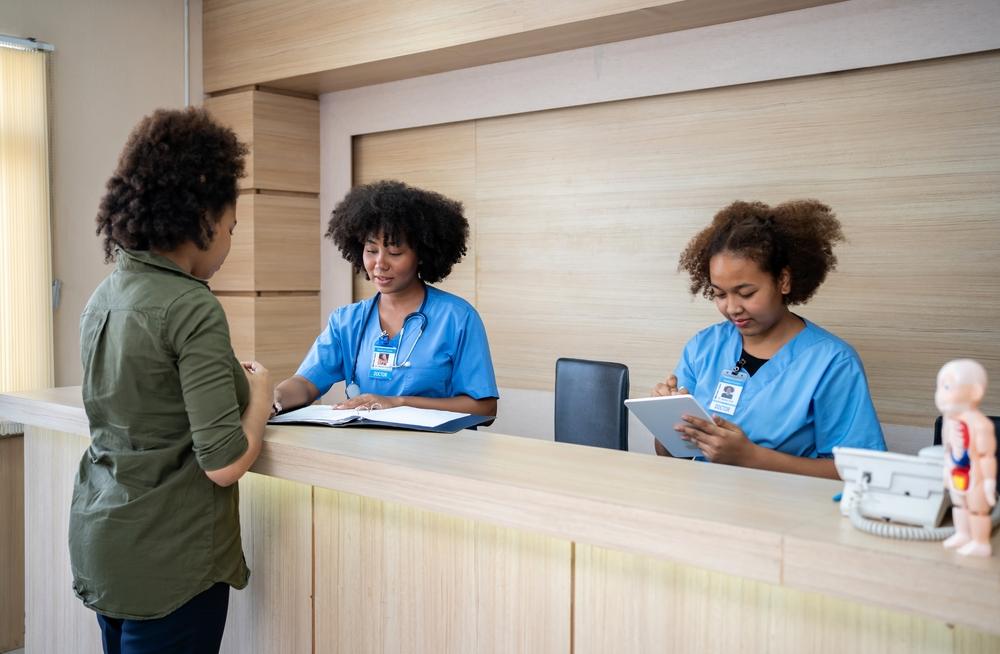 Paciente sendo atendida na recepção de um pronto-socorro por duas profissionais de saúde, representando cobertura de plano de saúde, atendimento de urgência e direito ao pronto-socorro.