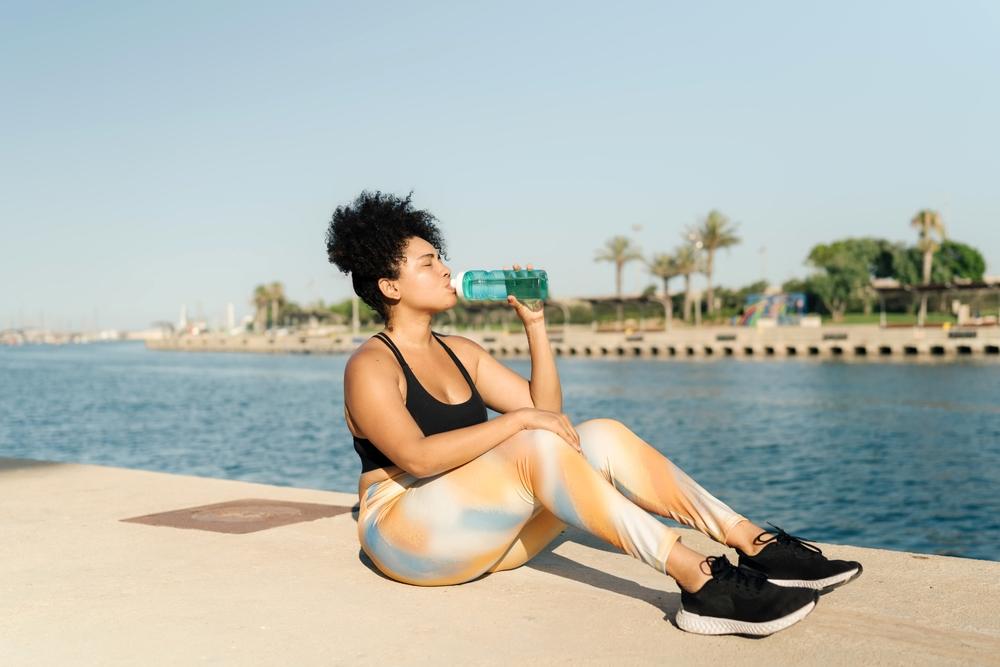 Prevenção da Insuficiência Renal. Pessoa sentada à beira de um cais, vestindo roupa esportiva e bebendo água após atividade física ao ar livre, destacando a importância da hidratação para a saúde dos rins.