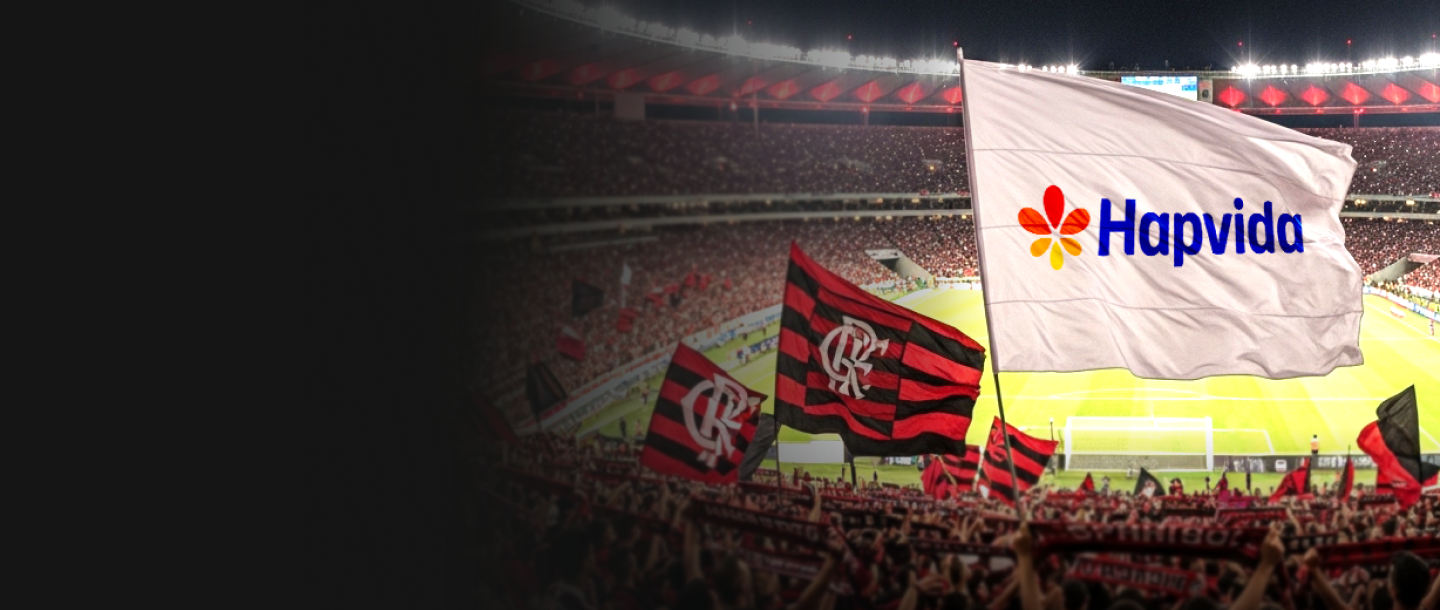 Bandeira da Hapvida sendo exibida em estádio de futebol lotado, com torcedores e bandeiras rubro-negras ao fundo, representando apoio ao esporte e à saúde.